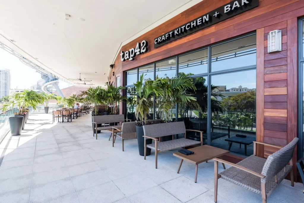 Covered patio with woven lounge chairs and potted palms outside the wood-clad storefront of Tap 42 Aventura