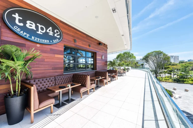 Tufted leather booths lining the sunny outdoor terrace beneath the oval logo sign at Tap 42 Aventura