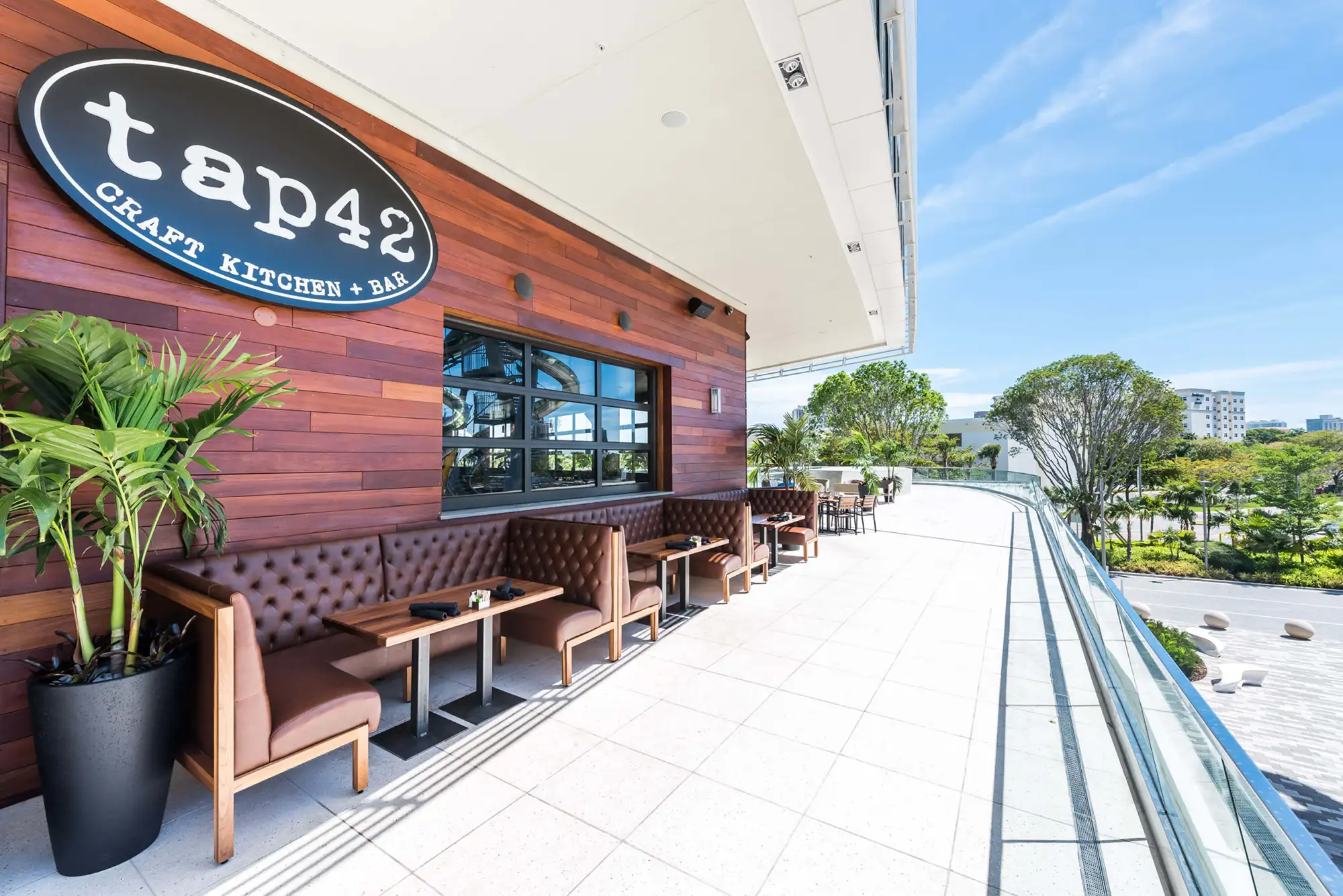 Tufted leather booths lining the sunny outdoor terrace beneath the oval logo sign at Tap 42 Aventura