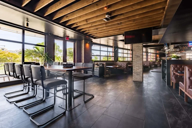 High-top tables with gray leather chairs by tall windows under a wood-beam ceiling at Tap 42 Aventura