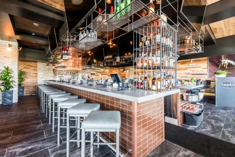 Copper-tiled bar with white stools and liquor bottles in hanging wire racks at Tap 42 Aventura