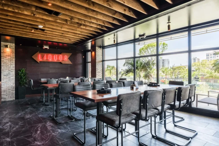 High-top tables by floor-to-ceiling windows facing a red neon liquor arrow sign at Tap 42 Aventura
