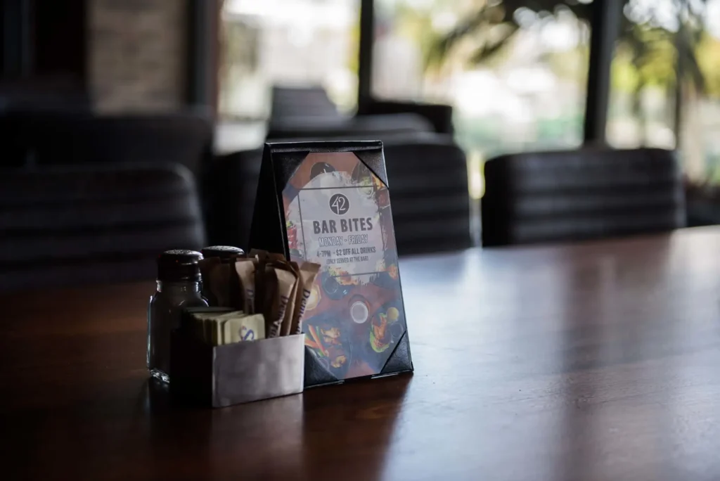 Bar bites happy hour table card beside salt and sugar packets on a wood table at Tap 42 Aventura