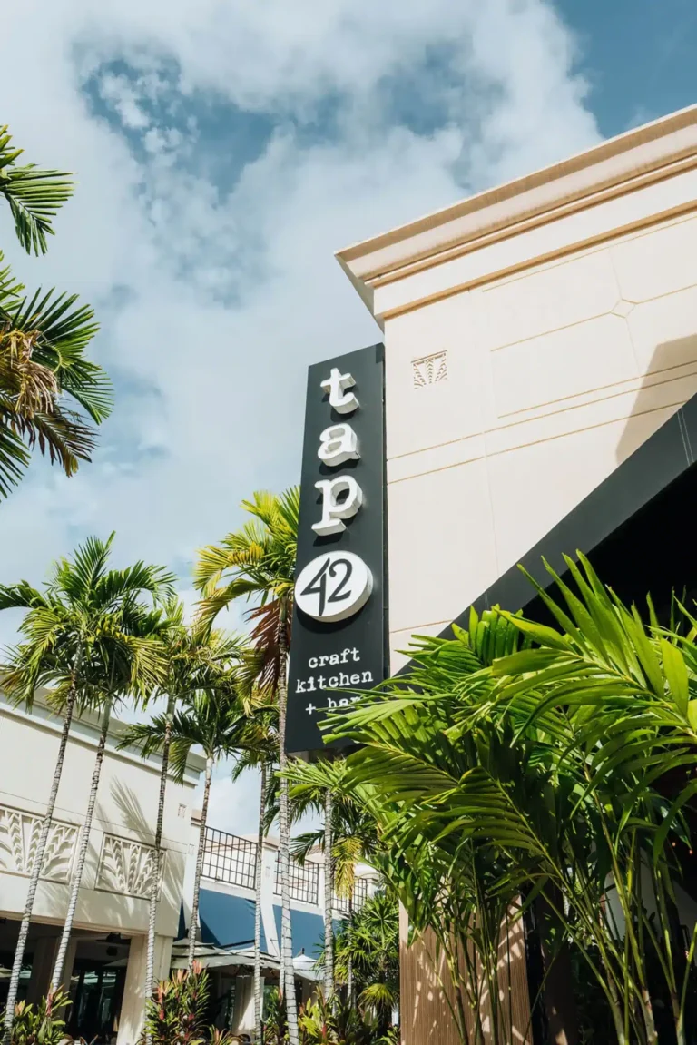 Vertical black sign reading tap 42 craft kitchen + bar on a cream facade framed by palm trees at Tap 42 Boca Raton