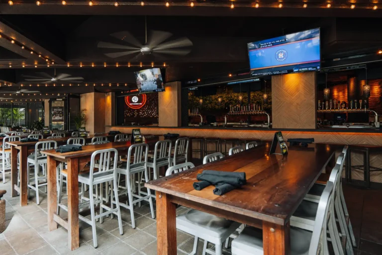 Wood high-top tables with aluminum chairs under string lights and TVs on the covered patio bar at Tap 42 Boca Raton
