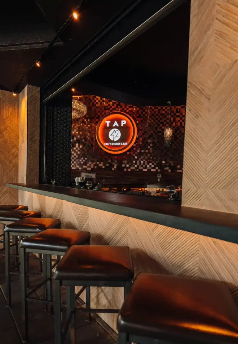 Leather stools at a herringbone wood bar counter, glowing Tap 42 logo on a mosaic tile wall beyond, at Tap 42 Boca Raton