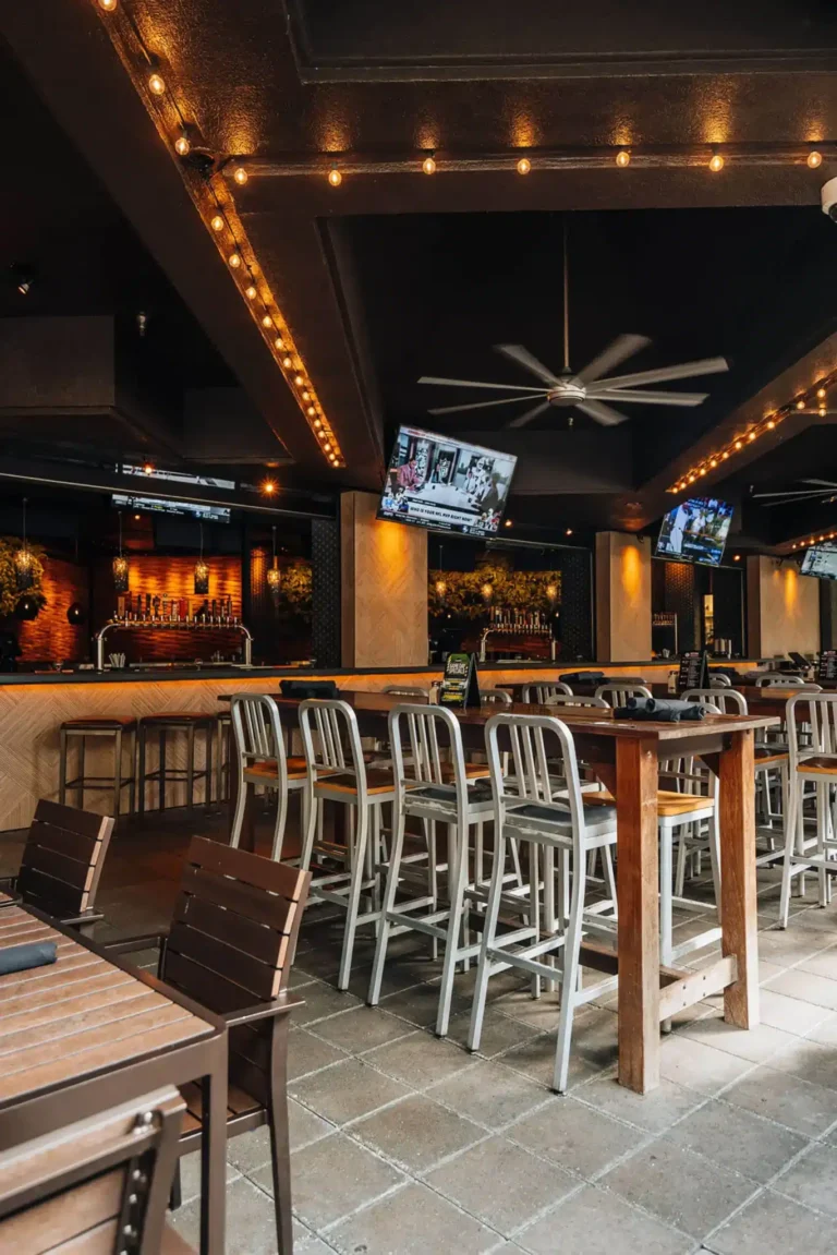 Patio high-tops with aluminum stools beneath a ceiling fan, string lights and TVs facing the bar at Tap 42 Boca Raton