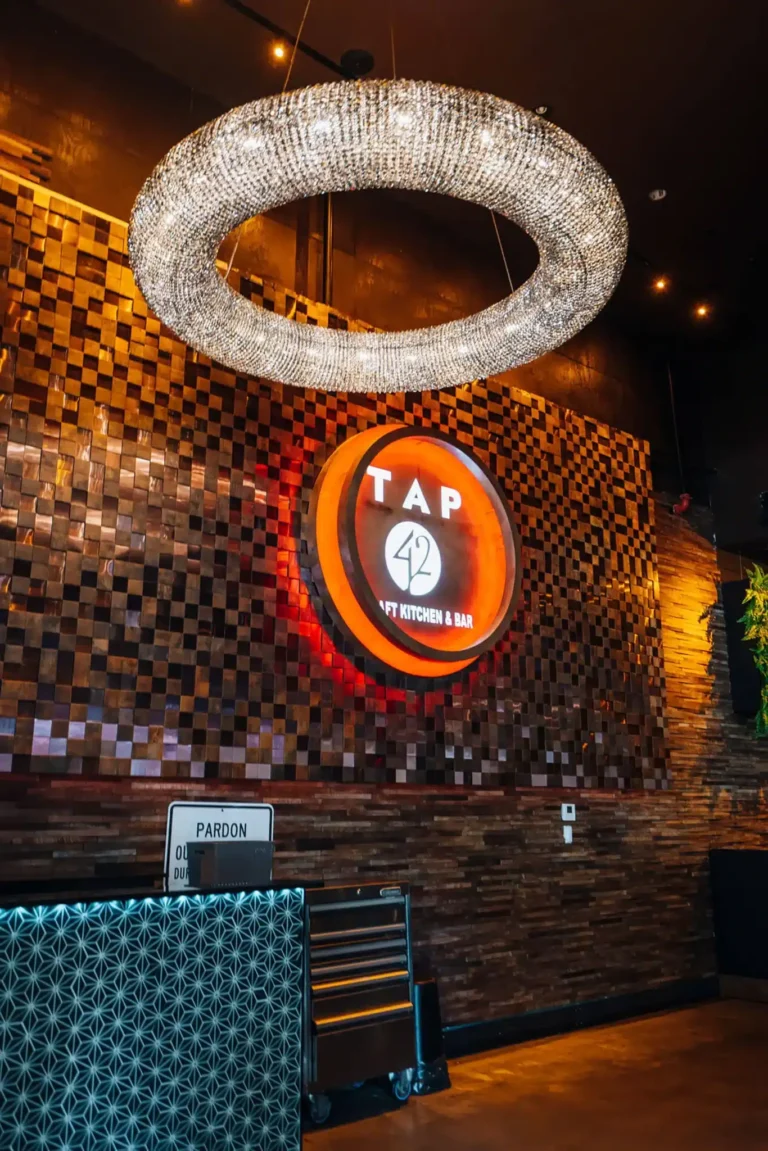 Ring-shaped crystal chandelier above the illuminated Tap 42 sign and host stand on a mosaic wall at Tap 42 Boca Raton