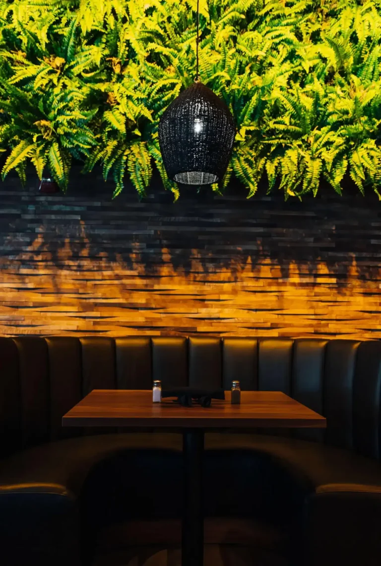 Single booth table beneath a woven pendant light and lush fern wall over warm-lit stacked wood at Tap 42 Boca Raton