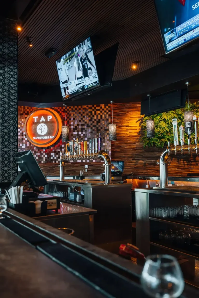 Beer tap towers behind the bar beneath the glowing Tap 42 logo on a mosaic tile wall with TVs above at Tap 42 Boca Raton