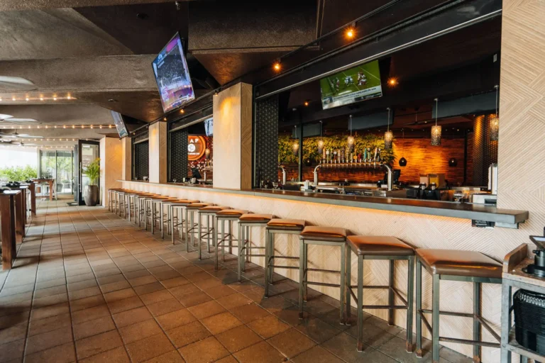 Long row of leather stools along the herringbone bar on a tiled walkway with TVs and string lights at Tap 42 Boca Raton