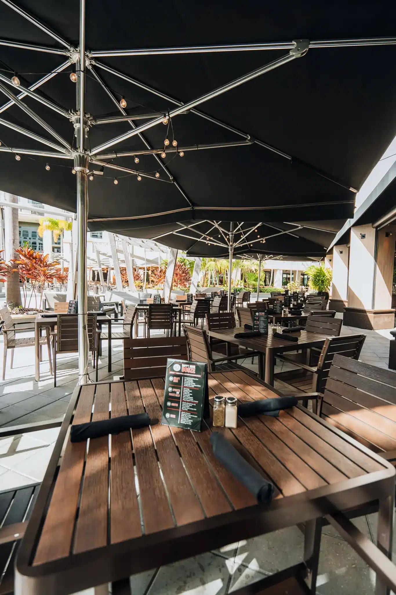 Sunny patio tables shaded by large black umbrellas strung with lights, tropical plants beyond, at Tap 42 Boca Raton