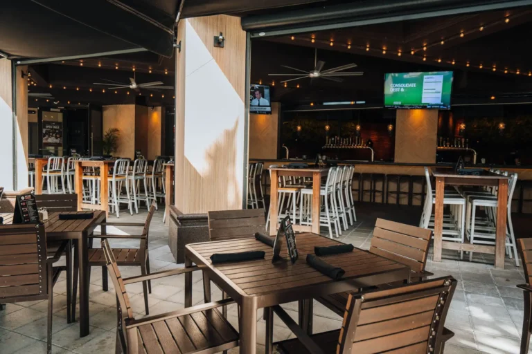 Patio dining tables in front of a covered bar area with white high-top chairs, string lights and TVs at Tap 42 Boca Raton