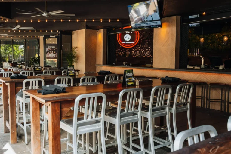 Communal high-tops with aluminum chairs facing the bar and a glowing Tap 42 logo on a mosaic wall at Tap 42 Boca Raton