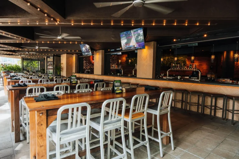 Communal high-tops beside empty bar stools at the open-air bar, daylight at the far end, at Tap 42 Boca Raton