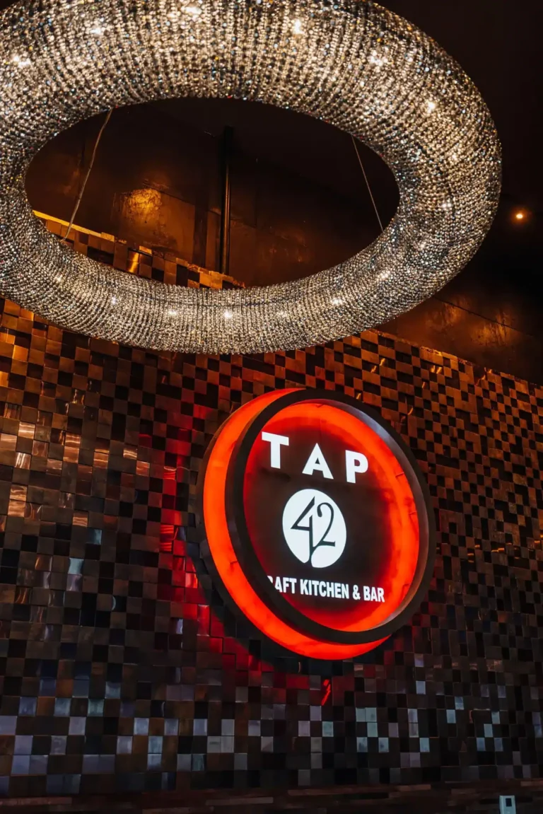 Close-up of the ring crystal chandelier hanging above the glowing red Tap 42 sign on mosaic tile at Tap 42 Boca Raton