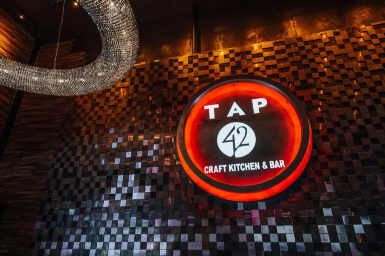Low-angle view of the illuminated Tap 42 Craft Kitchen & Bar sign on a mosaic tile wall at Tap 42 Boca Raton