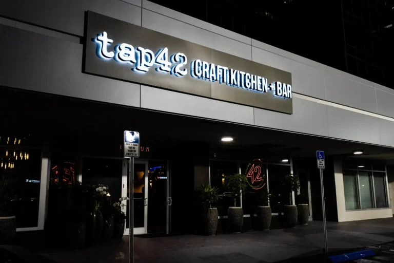 Entrance at night with backlit sign, potted plants and a neon 42 in the window at Tap 42 Coral Springs