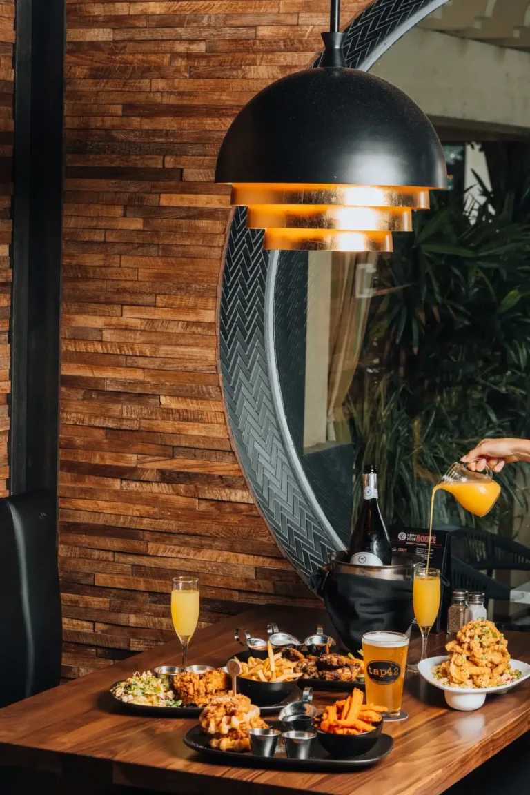 Brunch spread with chicken and waffles, fries and mimosas as juice is poured beside a round window at Tap 42 Naples