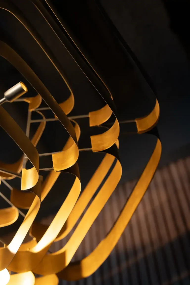 Close-up of a sculptural gold ribbon pendant light glowing against a dark ceiling at Tap 42 Naples