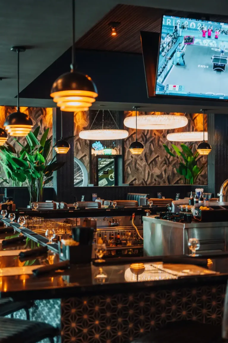 Bar counter with glassware and dome pendant lights below a TV, carved wall reflected in a mirror, at Tap 42 Naples
