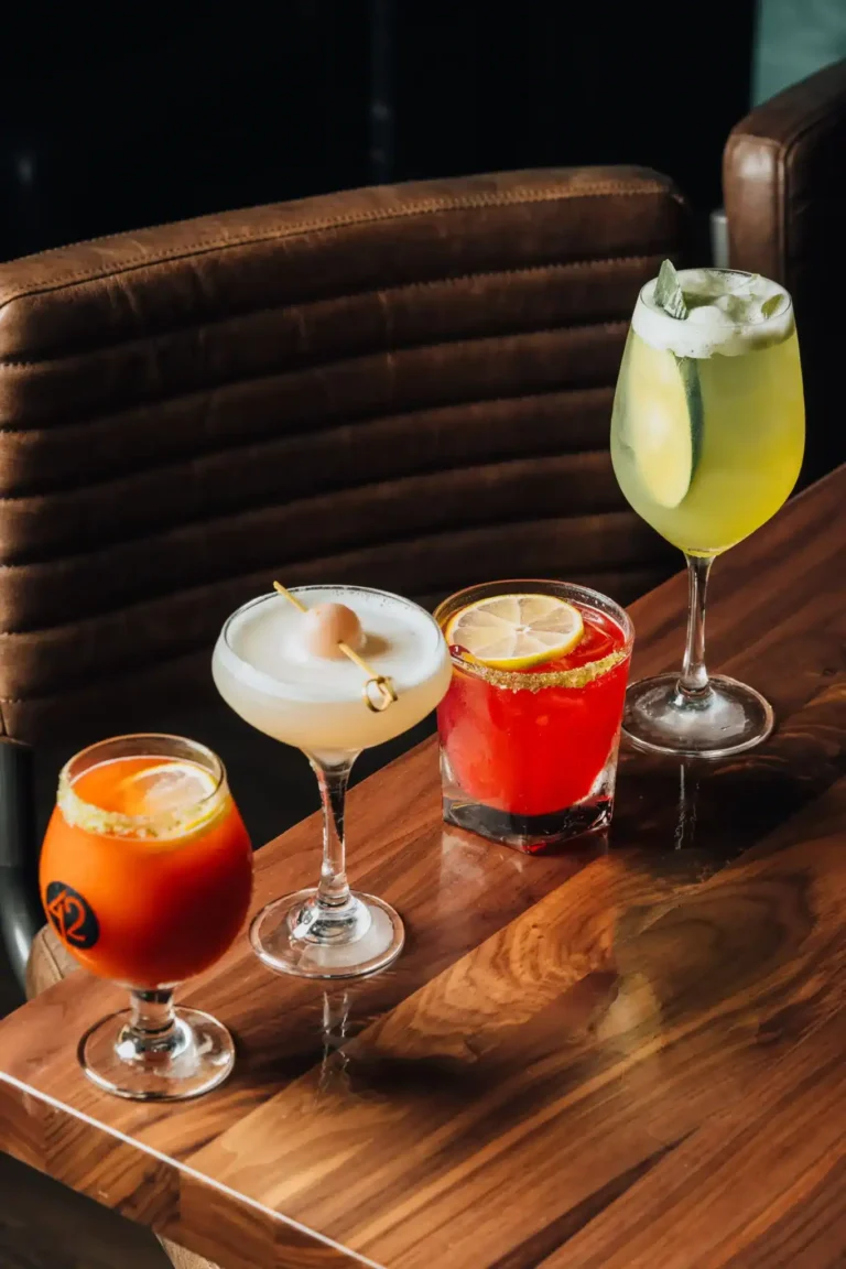 Four colorful cocktails, from a salt-rimmed red drink to a cucumber spritz, on a wood table at Tap 42 Naples
