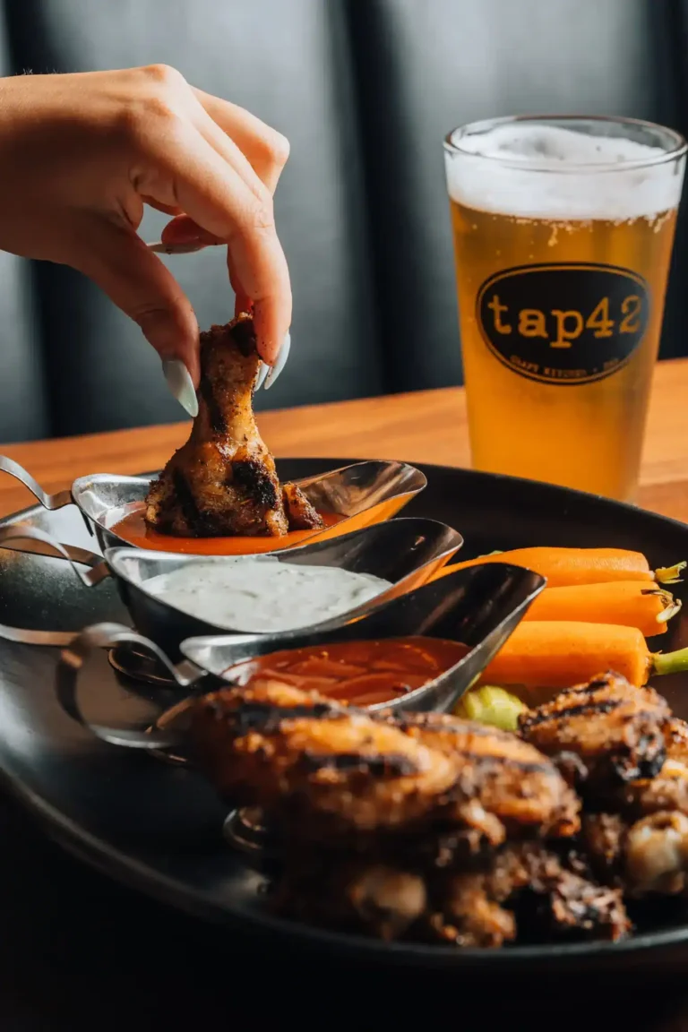 Guest dipping a grilled chicken wing into sauce beside ranch, carrots and a branded beer at Tap 42 Naples