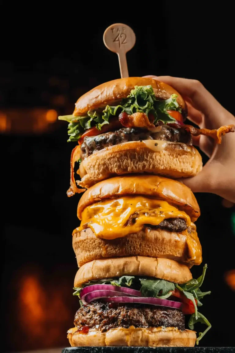 Hand holding a towering stack of three loaded burgers topped with a logo pick at Tap 42 Naples