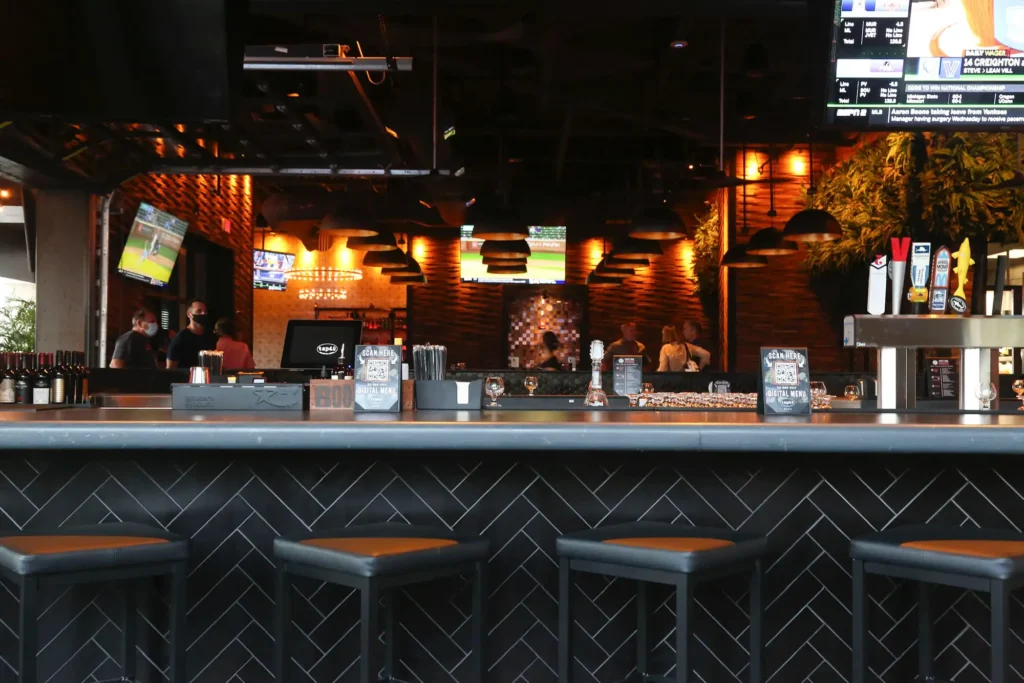 Bar stools at a black herringbone-tiled bar with beer taps, dome pendant lights and TVs at Tap 42 Davie