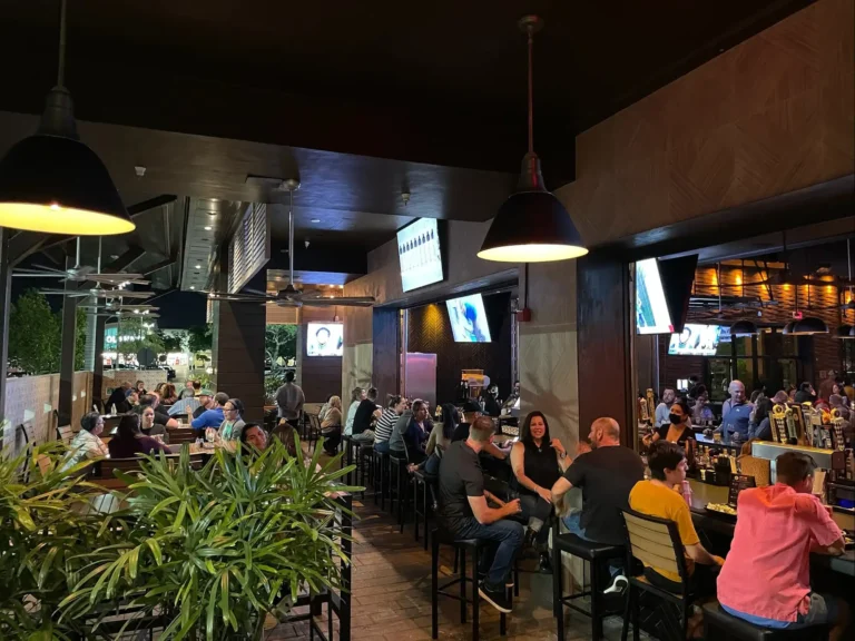 Guests at high-top bar seating along the open-air patio bar at night under black pendant lights at Tap 42 Davie