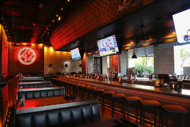 Empty leather booths and bar stools under a glowing red neon 42 sign and TVs before opening at Tap 42 Davie