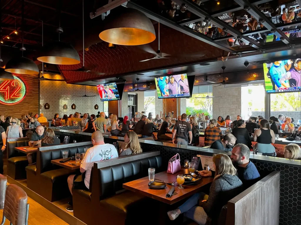 Crowded dining room with guests in booths sharing drinks and food while TVs show sports at Tap 42 Davie