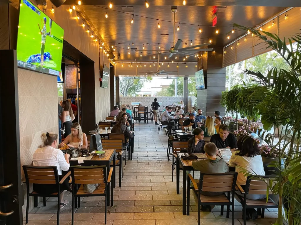 Guests dining at patio tables under string lights and ceiling fans with a TV and potted palms at Tap 42 Davie