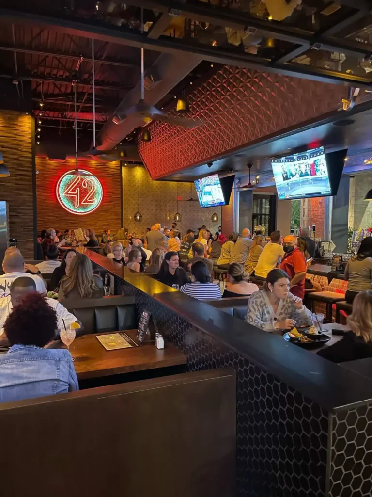 Guests at booths and the bar in a vertical view toward the red neon 42 sign and TVs at Tap 42 Davie
