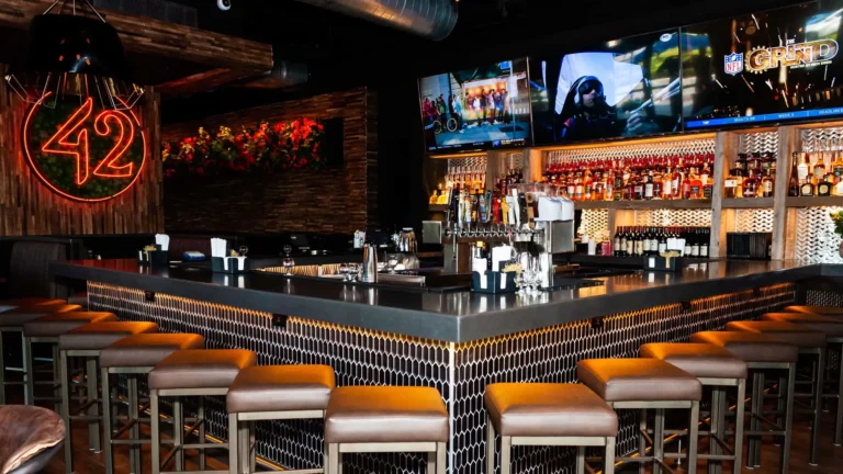 Square bar with leather stools, hex-tile front, liquor shelves and TVs beside a neon 42 sign at Tap 42 Doral