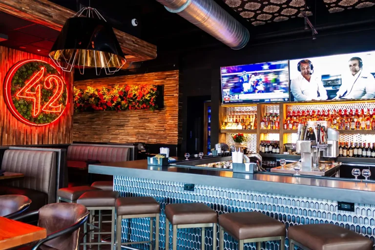 Bar corner with blue hexagon tile, brown leather stools and wall TVs near the succulent neon 42 at Tap 42 Doral