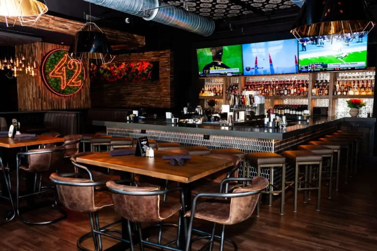 Dimly lit dining room with Edison bulb chandeliers, high-top tables and the bar beyond at Tap 42 Doral