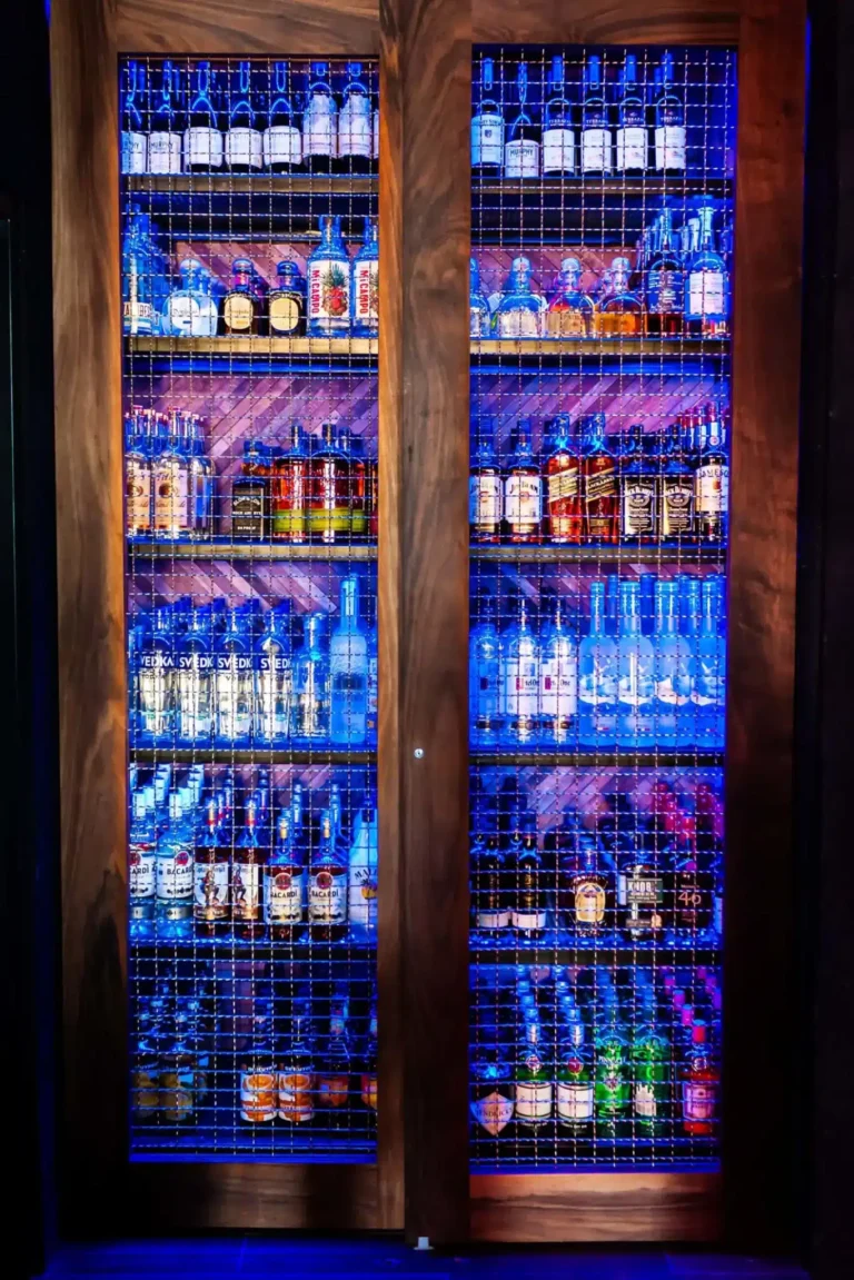 Blue-lit liquor cabinet with wire mesh doors stocked with vodka, rum and whiskey bottles at Tap 42 Doral