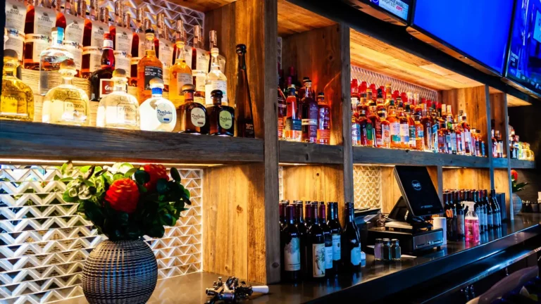 Back bar shelves of tequila and wine bottles with a red flower vase and POS screen at Tap 42 Doral