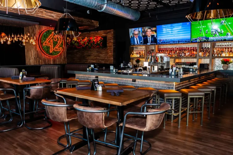 Warm-lit view of high-top dining tables and the full bar with sports TVs above at Tap 42 Doral