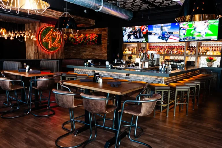 Lower-angle view across a leather-chair high-top toward the bar and neon 42 sign at Tap 42 Doral