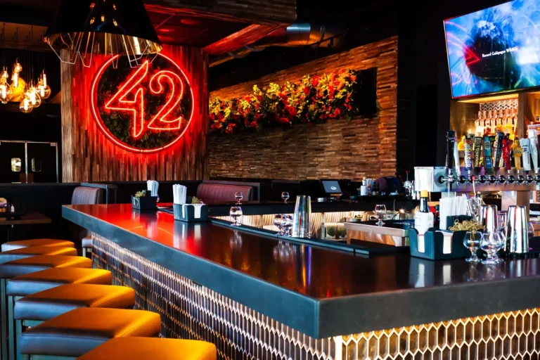 Amber-lit bar stools along a glossy bar top with beer taps and the neon 42 beyond at Tap 42 Doral
