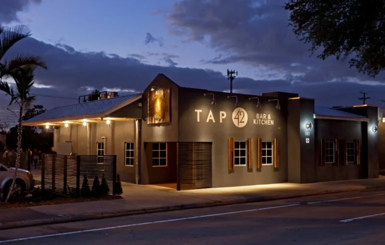 Gray building with a glowing Bar & Kitchen sign and wood shutters at dusk at Tap 42 Fort Lauderdale