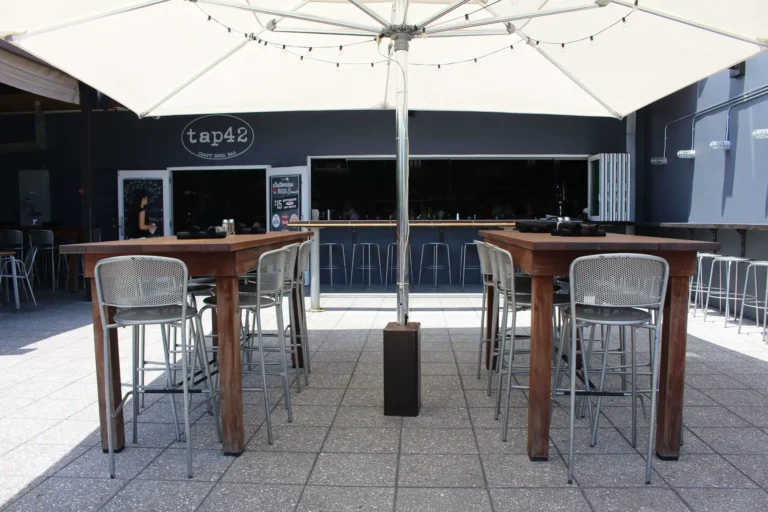 Sunny patio with wood high-top tables under a white umbrella at Tap 42 Fort Lauderdale