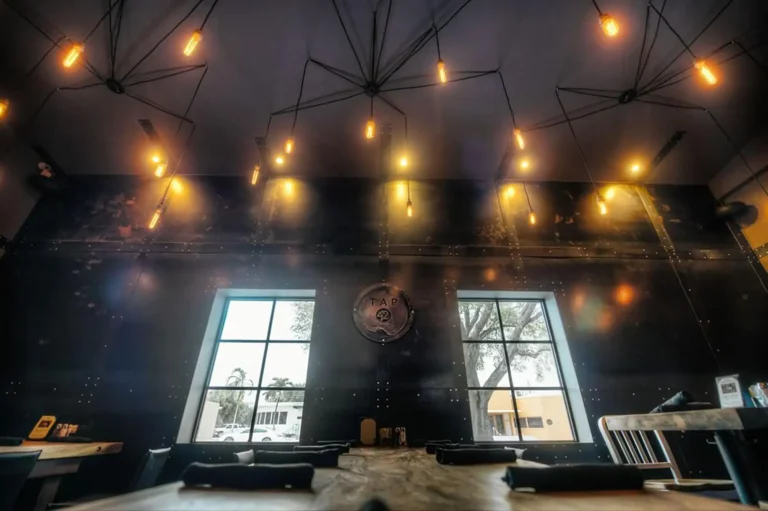 Edison bulb chandeliers above a dark riveted wall and tall windows at Tap 42 Fort Lauderdale