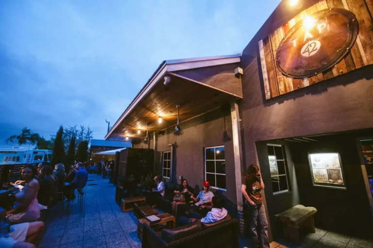 Guests on lounge sofas and patio tables at dusk beneath a round wood sign at Tap 42 Fort Lauderdale