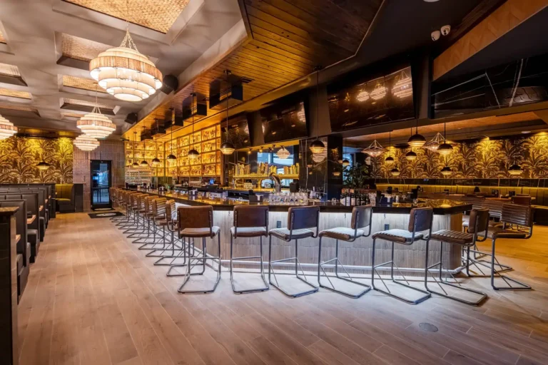 Wide view of the long bar, tiered chandeliers and palm-print wallpaper in the dining room at Tap 42 Fort Myers