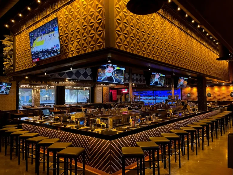 Center bar with stools and sports TVs under gold geometric walls, windows beyond at Tap 42 Kendall