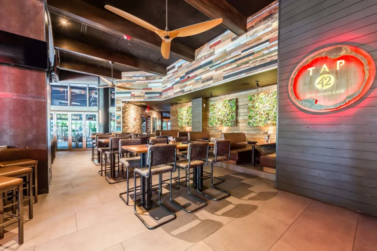 Dining room with high-tops, ceiling fan, plant wall panels and glowing logo sign at Tap 42 Midtown Miami