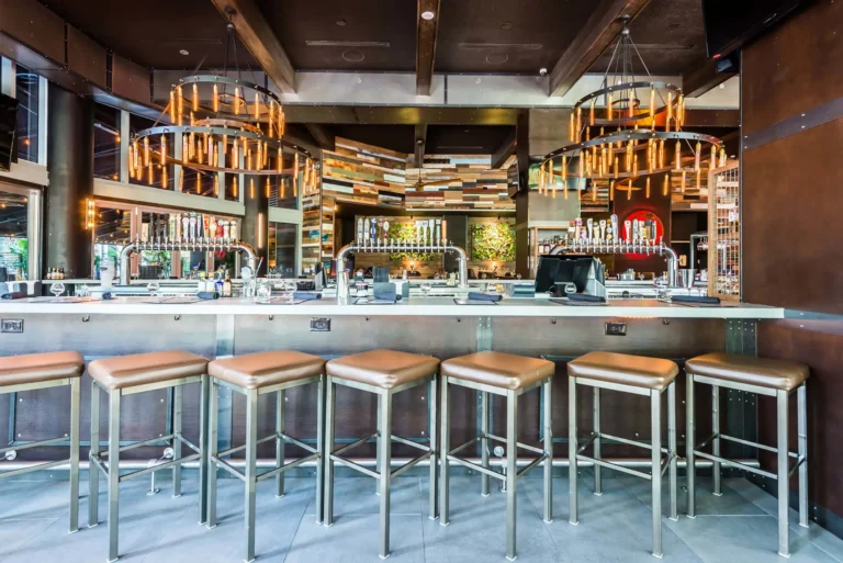 Front view of the bar with tan stools, beer tap towers and two Edison bulb chandeliers at Tap 42 Midtown Miami